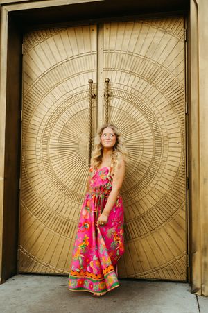 Young woman in vibrant pink floral maxi dress standing before ornate gold circular patterned double doors.