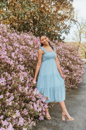 Woman in light blue tiered midi dress standing near pink flowering bushes outdoors.