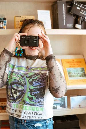 Young girl holding a vintage black film camera up to her face, wearing a graphic tee in a camera shop.
