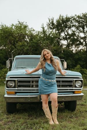 Smiling blonde woman in blue denim outfit and tan boots posing in front of a vintage Ford truck.