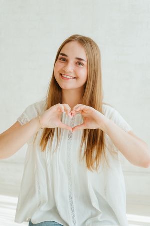 Smiling young woman with long brown hair making a heart shape with her hands wearing a white blouse.