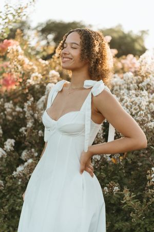 Woman in white tie-strap sundress smiling among blooming garden flowers at sunset.
