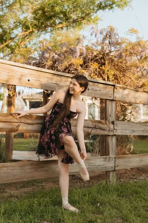 Young woman in floral dress posing gracefully by a rustic wooden fence with blooming wisteria.