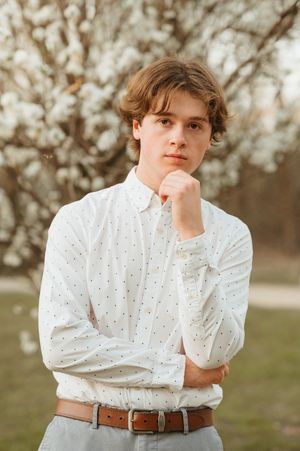 Young man in white polka dot shirt posing thoughtfully near blooming trees.