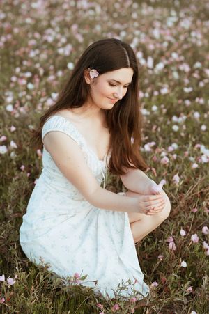 Young woman in light floral dress sitting among pink wildflowers with butterfly hair clip.