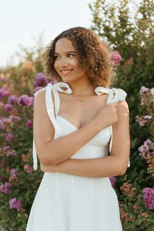 Curly-haired woman in white sundress smiling among pink and purple garden flowers.