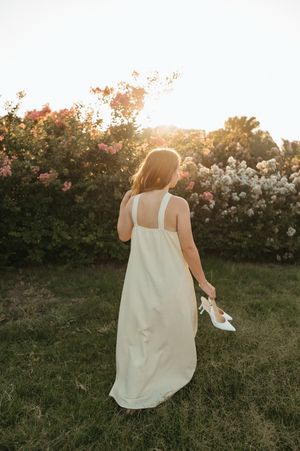 A woman in a cream maxi dress walks barefoot through a sunlit garden holding white heels.
