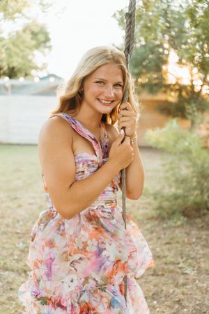 Young blonde woman in floral dress smiling while holding a rope swing outdoors at golden hour.