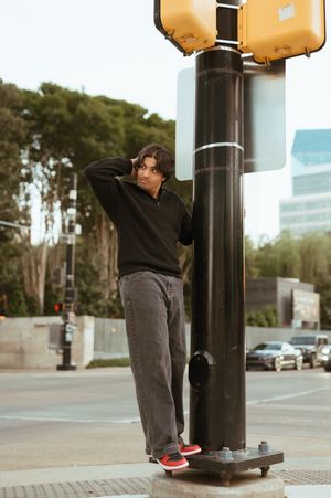 Young man in black hoodie and gray jeans leaning against a traffic light pole on a city street.