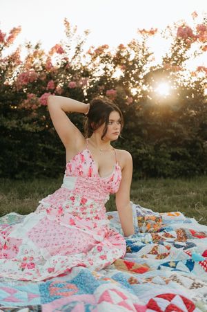 Young woman in pink floral dress sitting on colorful quilt at sunset with blooming trees.