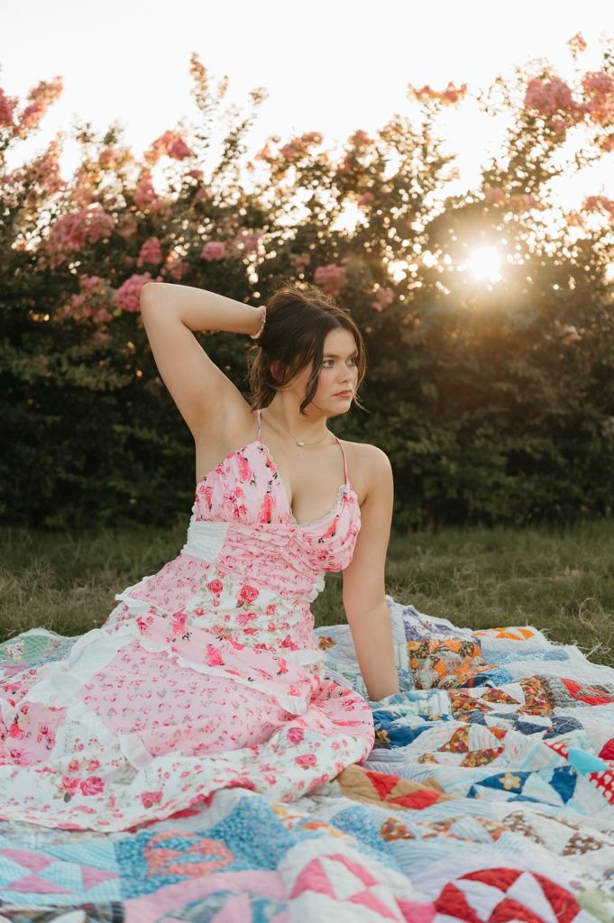 Young woman in pink floral dress sitting on colorful quilt at sunset with blooming trees.