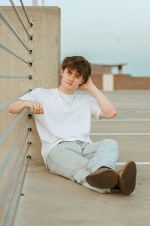 Young man in white t-shirt and light jeans sitting on parking garage floor leaning against wall.