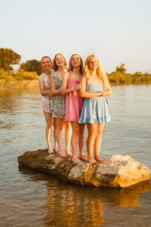 Four smiling teenage girls standing on a rock in a river, wearing summer dresses at golden hour.