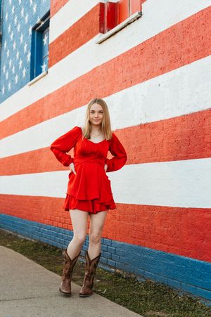 Young woman in red dress and cowboy boots posing by American flag mural.