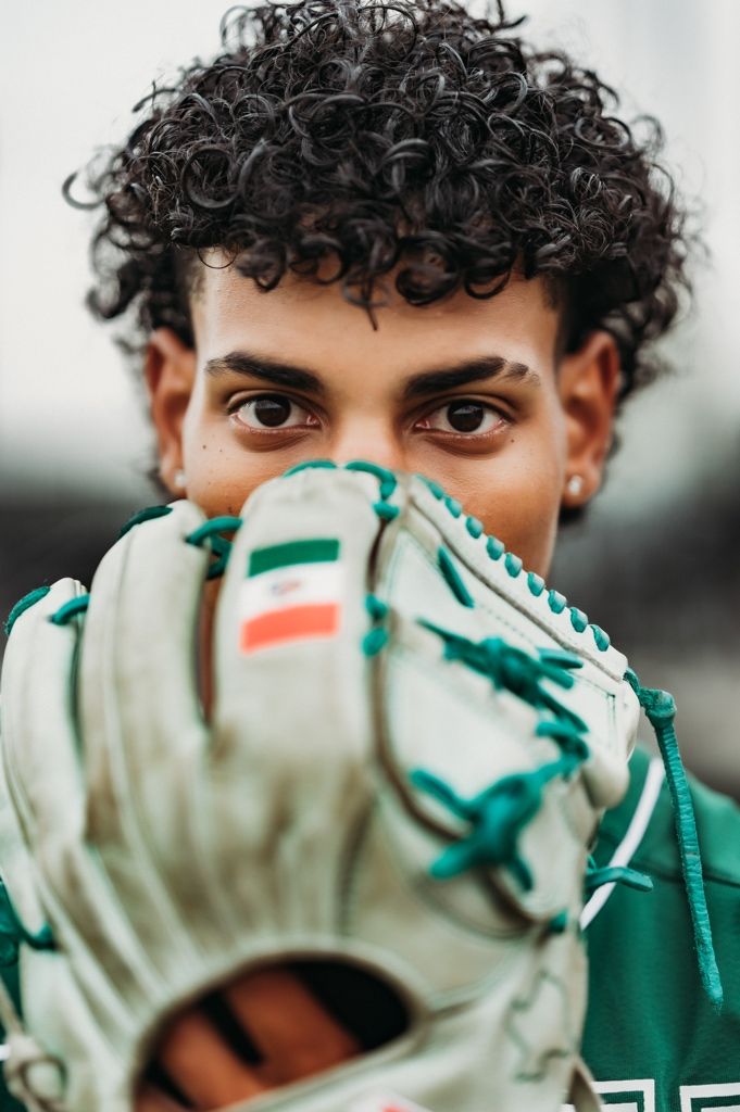 Young man with curly hair holding a green and white baseball glove up to his face outdoors.