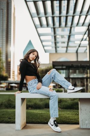 Young woman in black crop top, ripped jeans, and Nike sneakers sitting on a bench in an urban setting.