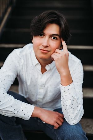 Young man in white polka dot shirt sitting on dark stairs, resting hand on temple in thoughtful pose.