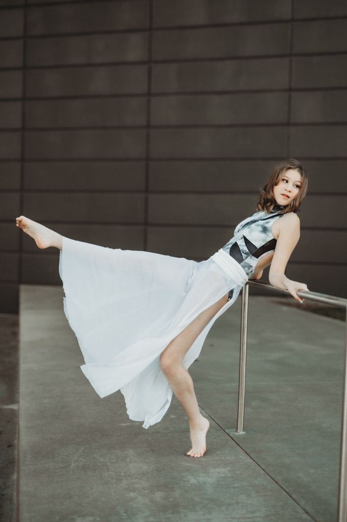 A barefoot woman in a flowing white skirt poses gracefully against a metal railing outdoors.