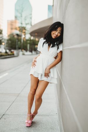 Young woman in white romper and pink heels leaning against wall on urban sidewalk, smiling.