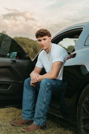 A young man in a white t-shirt and jeans sits casually in an open car door at sunset.