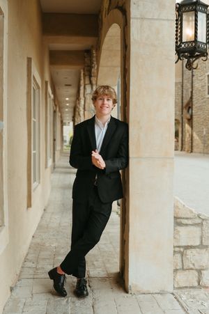Smiling teen in black suit leaning against stone column in outdoor corridor with lantern.