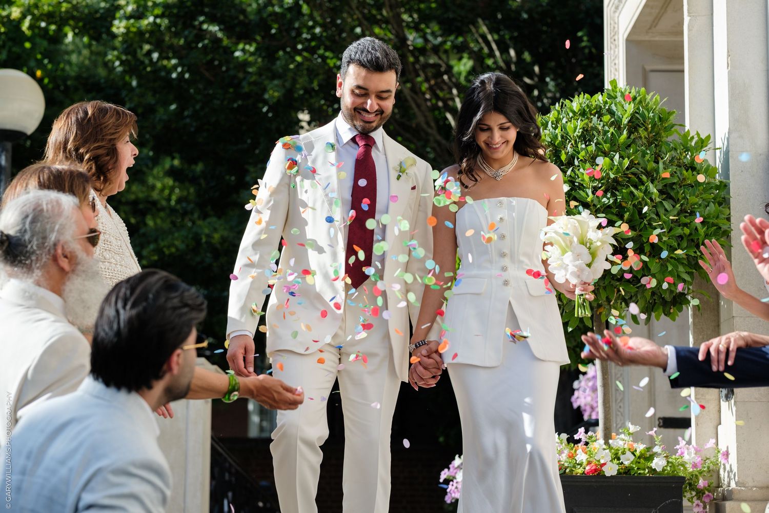 Newlywed couple in white suits holding hands while guests toss colorful confetti during outdoor wedding ceremony.