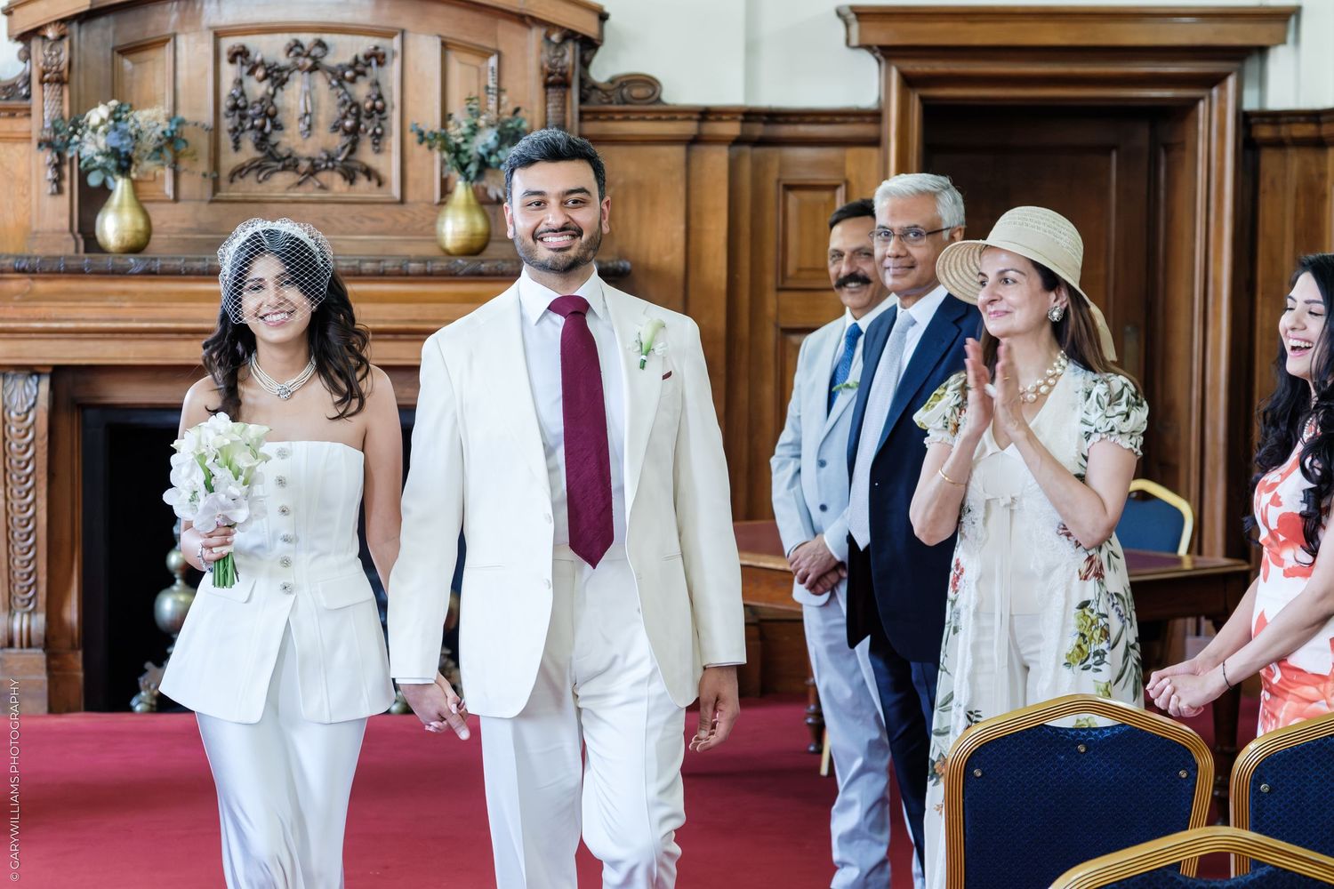 Bride and groom in white outfits walk hand-in-hand at a wedding ceremony while guests applaud in an elegant venue.
