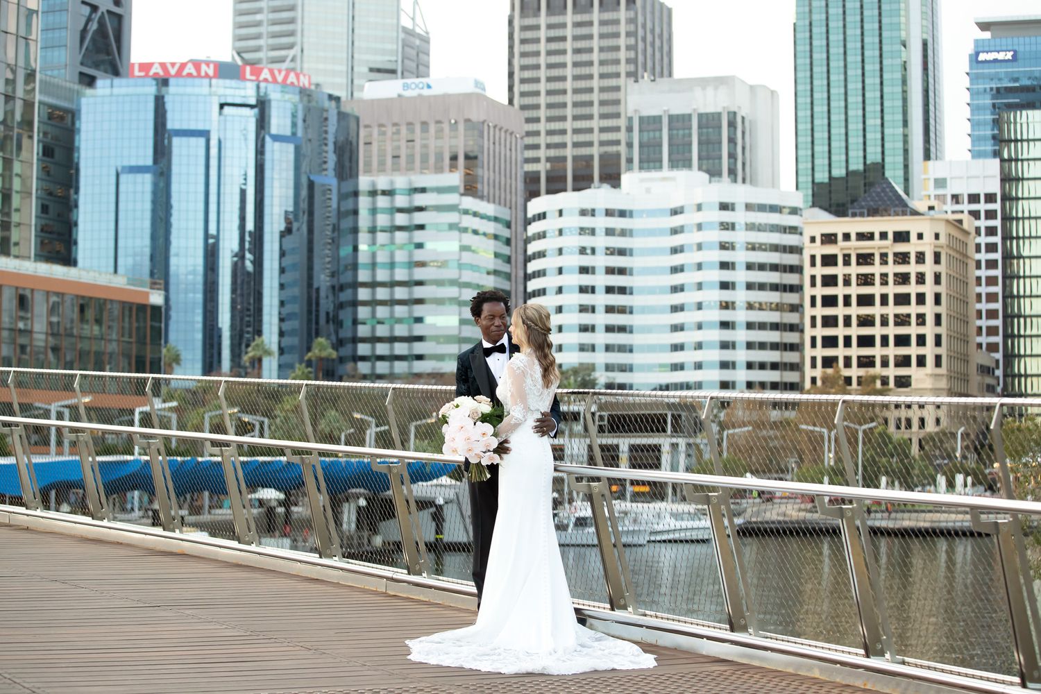 Bride and groom pose on a waterfront bridge with Sydney city skyline in the background on their wedding day.