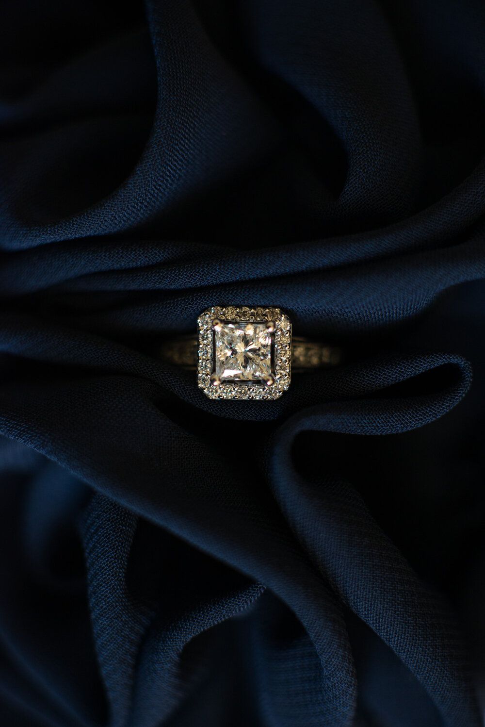 Gold halo engagement ring with princess-cut diamond displayed on dark navy blue silk fabric.
