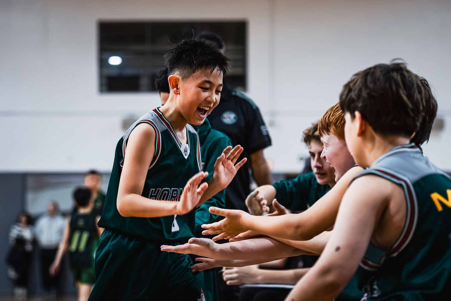 Young boy in green Hornets jersey dribbles basketball during indoor youth game, wearing red sneakers.