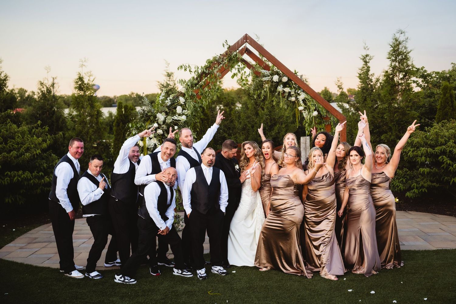 Wedding party celebrating outdoors under geometric arch, bridesmaids in gold gowns and groomsmen in black suits cheering at sunset.