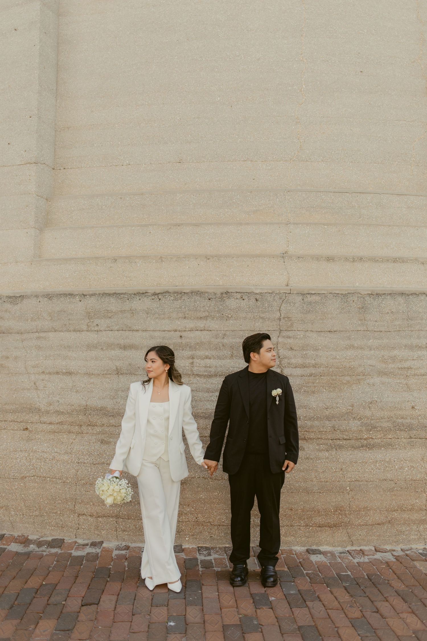 Bride in white suit and groom in black suit hold hands walking along a brick path against a stone wall.