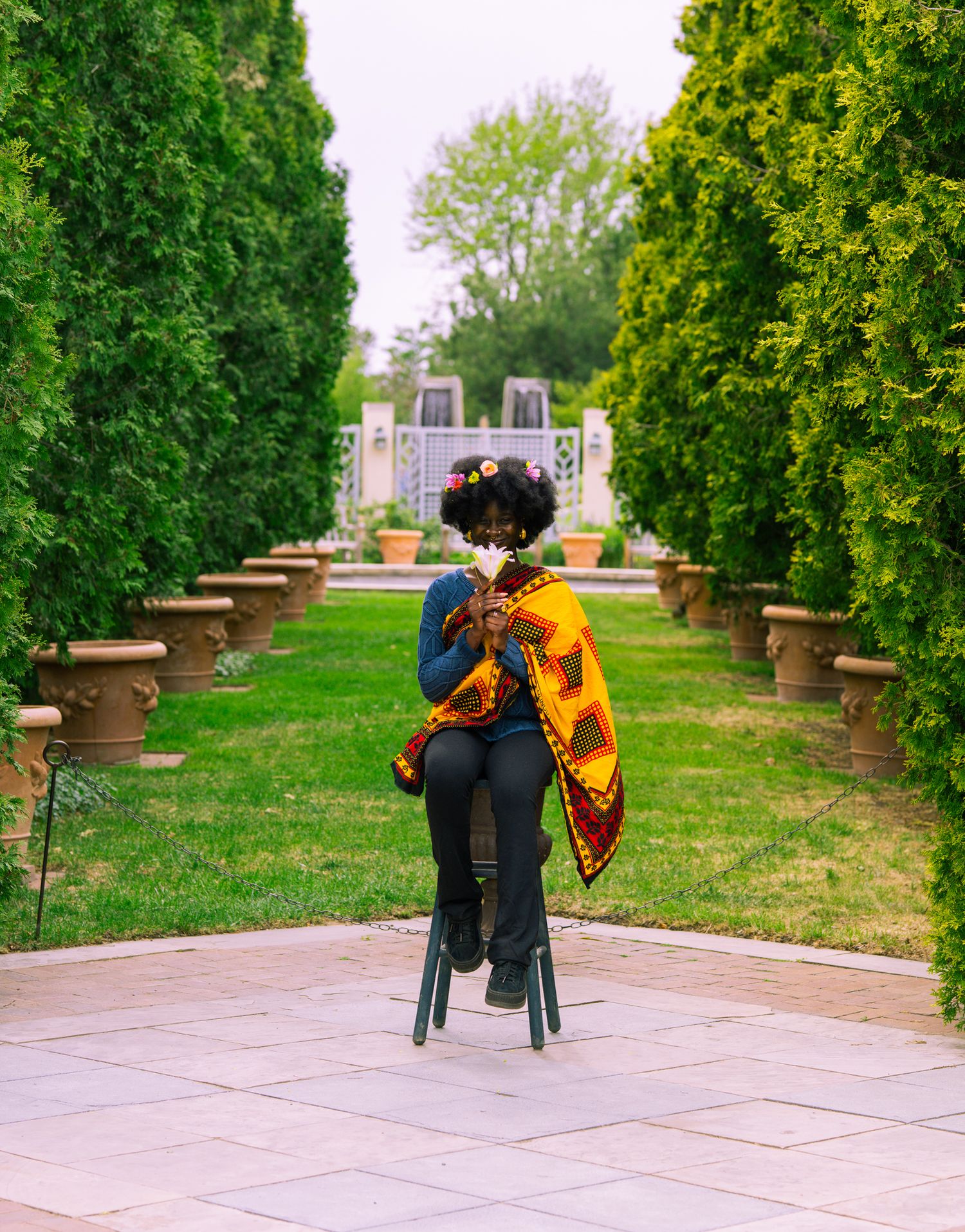 Person in colorful African print shawl sits on stool in lush garden pathway lined with trees and terracotta pots.