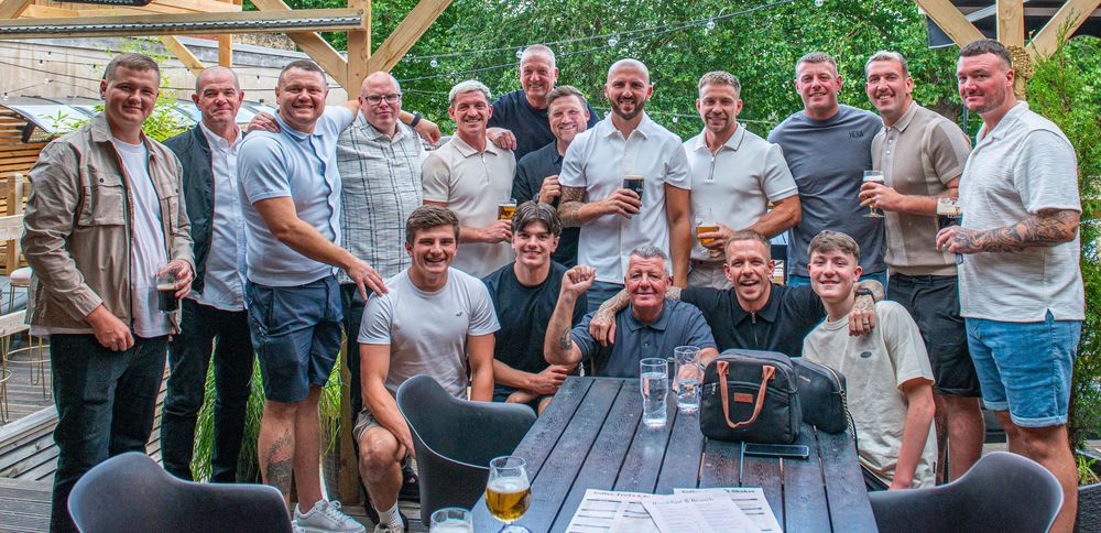 Large group of men posing together outdoors at a pub garden, smiling and holding drinks on a sunny day.