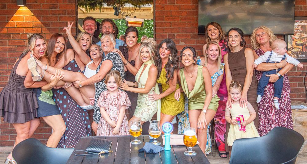 Large group of smiling women posing together at an outdoor bar patio, celebrating with drinks on a sunny day.