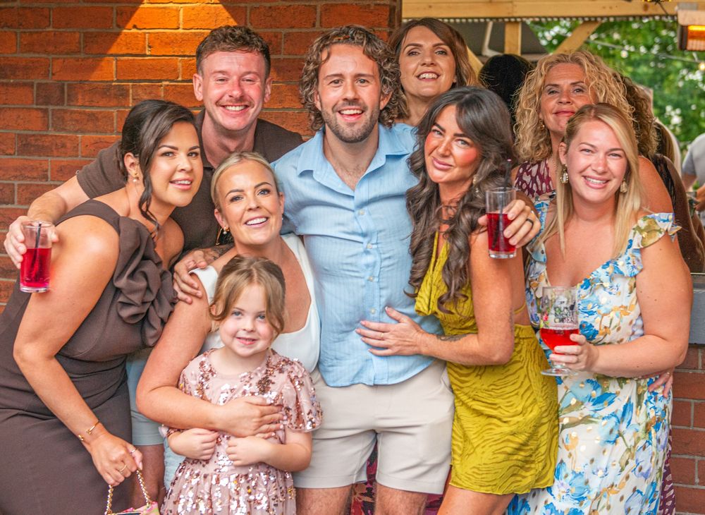 A joyful group of friends and family posing together outdoors at a summer gathering, smiling and holding drinks.