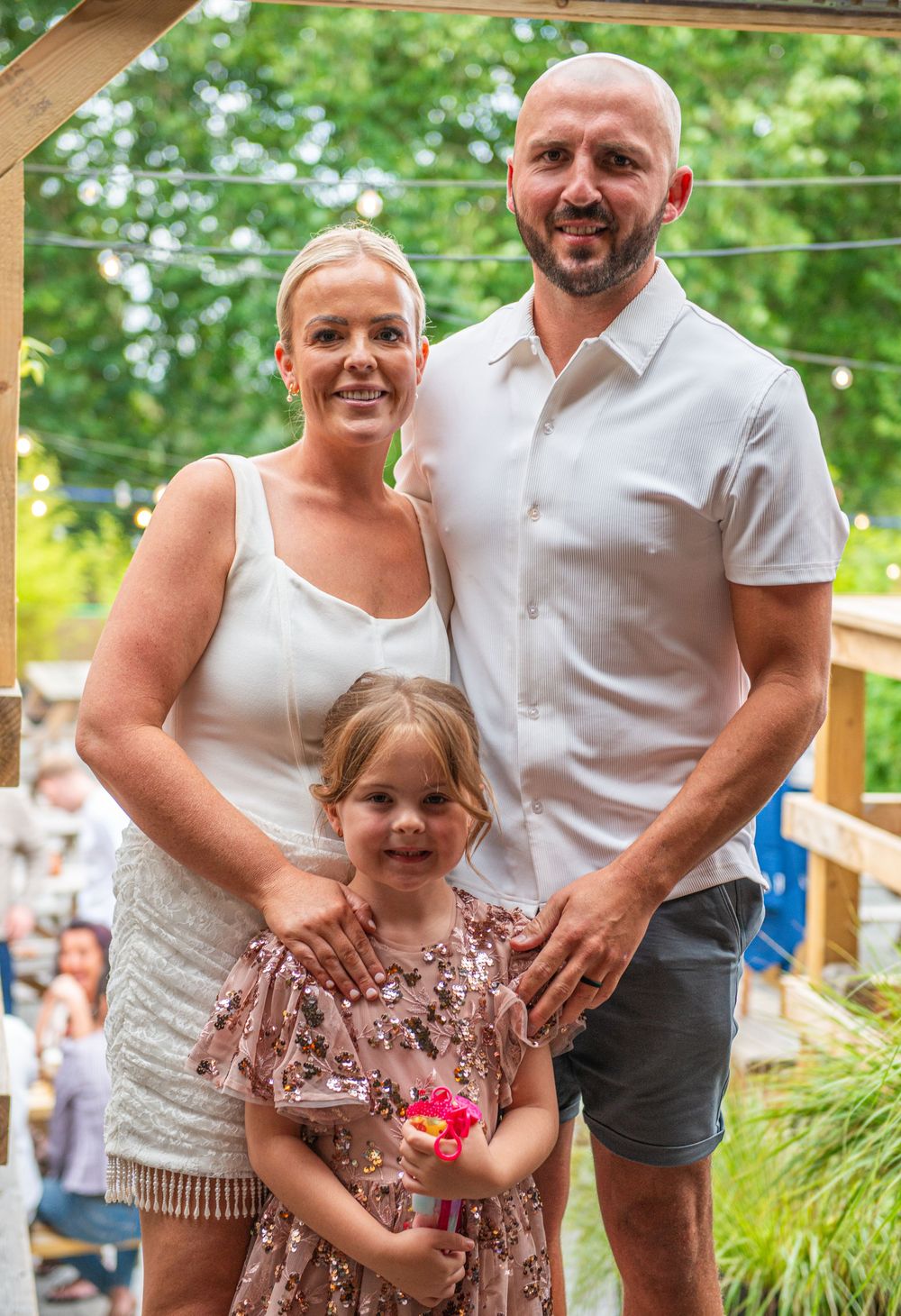 A smiling family of three poses outdoors, with two adults in white and a young girl in a sparkly pink dress.