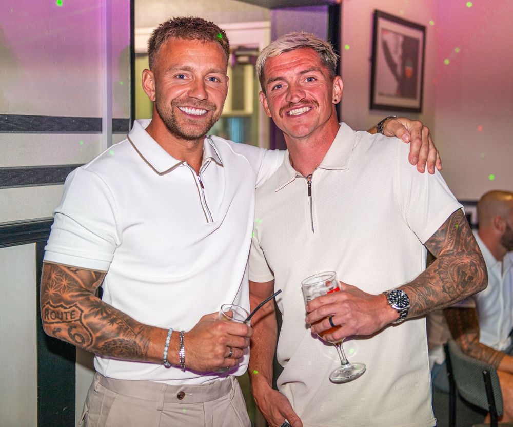 Two tattooed men in white polo shirts smiling and holding drinks at a social event.
