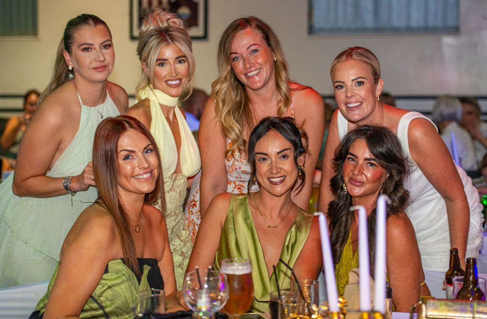 Seven women smiling and posing together at a festive dinner event, dressed in elegant attire.