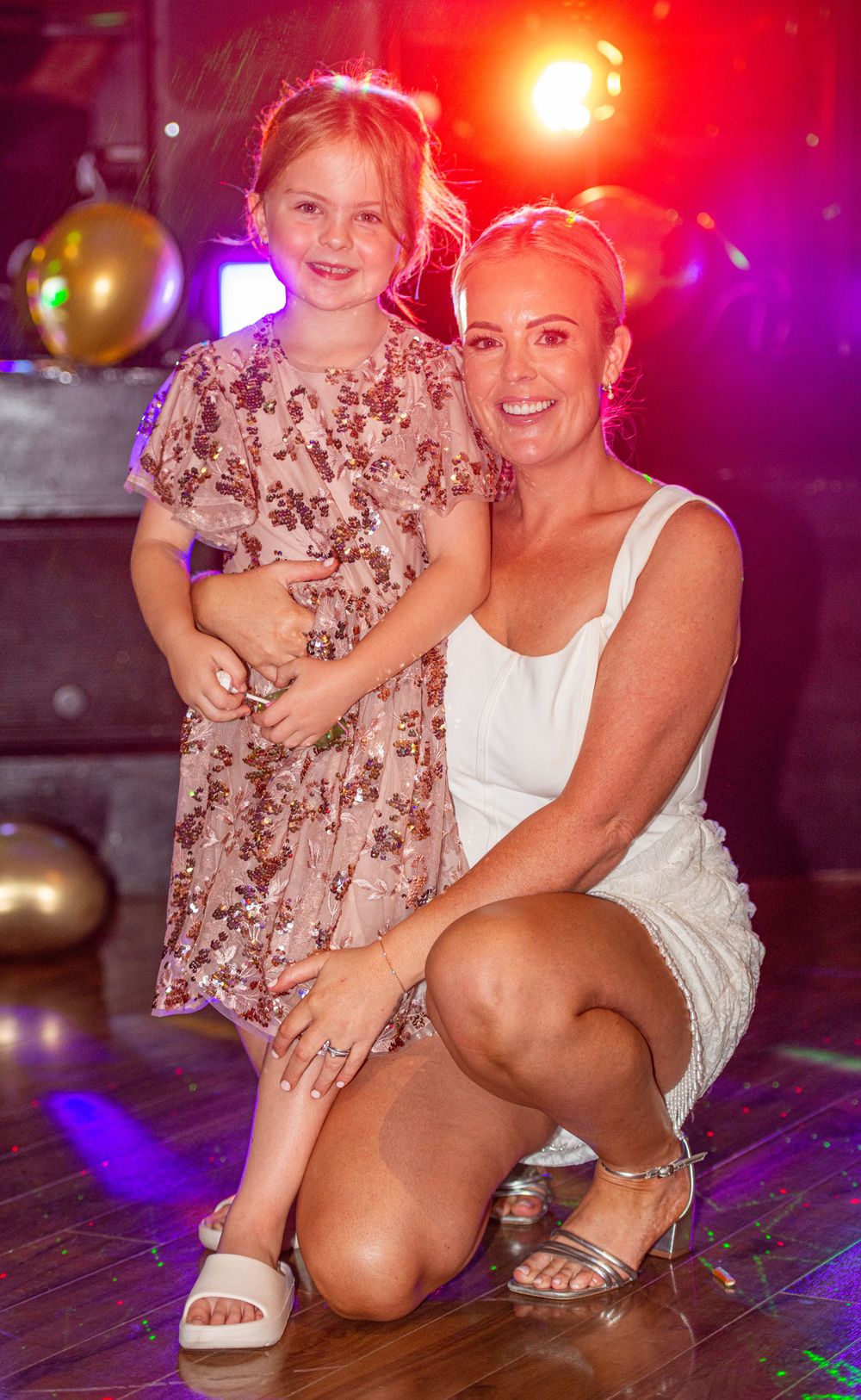 Woman in white dress crouches beside young girl in floral dress, posing together at a party with colorful stage lights.