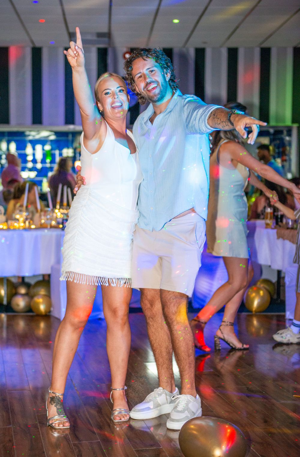 Couple in white outfits dancing and posing at a party event with colorful lights and balloons.