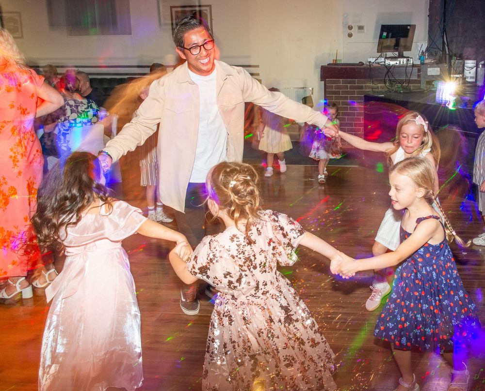 Adults and children holding hands dancing in a circle at a festive indoor party with colorful lights.