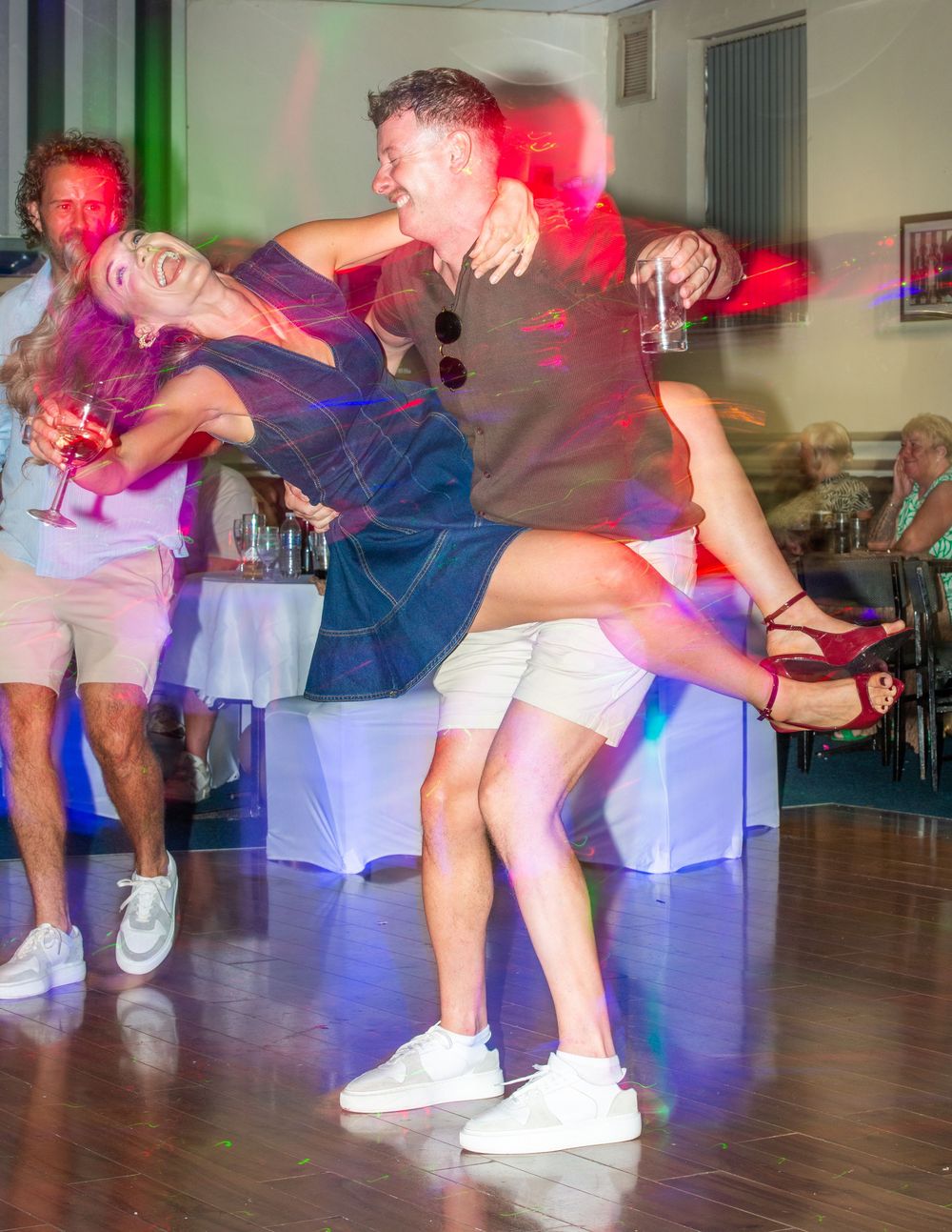 Couple dancing energetically on a dance floor at a party, with colorful lights and guests in the background.