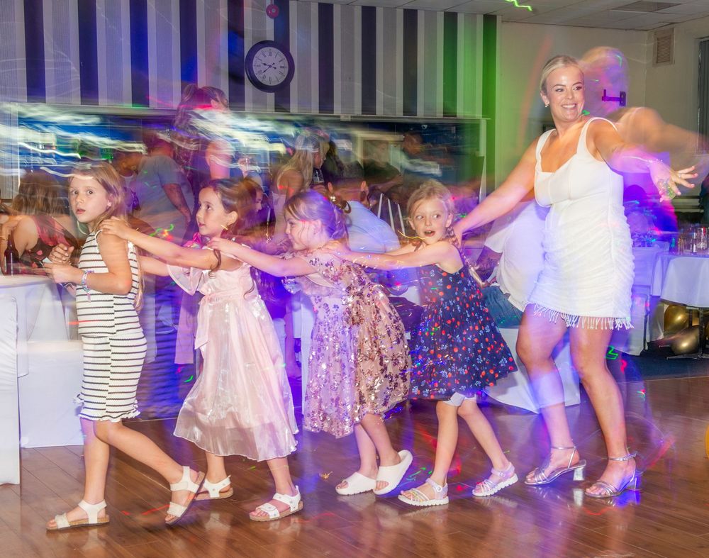 Women and girls dancing in a conga line at a party celebration on a colorful dance floor.