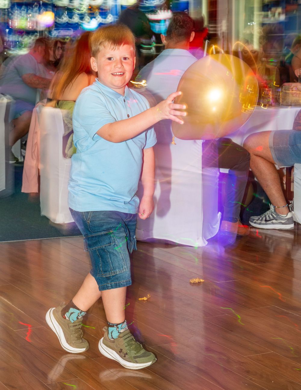 Young red-haired boy in blue shirt playing with gold balloon on dance floor at party celebration.