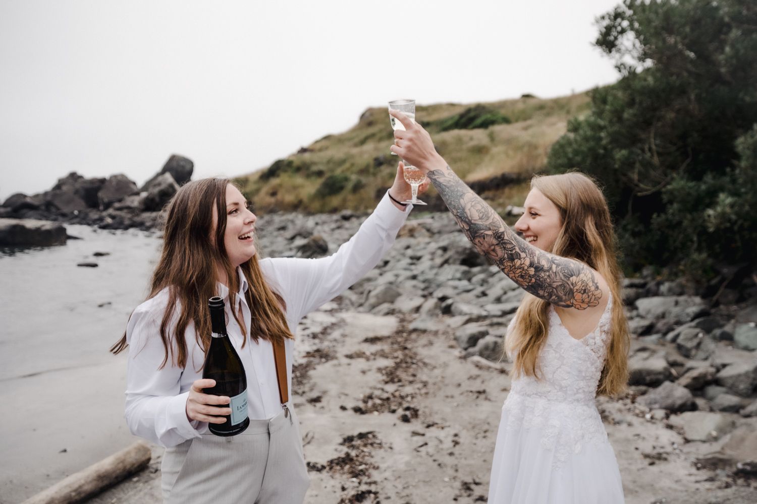 Two brides toast champagne glasses on the Oregon Coast after their elopement ceremony