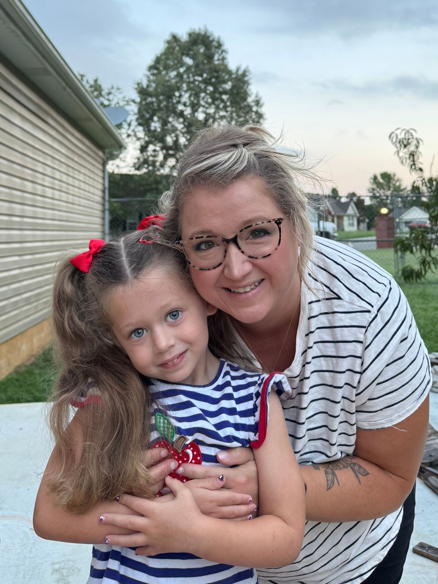 A mom with glasses hugs a young girl with red bows in her hair, both wearing striped shirts outdoors on first day of school.