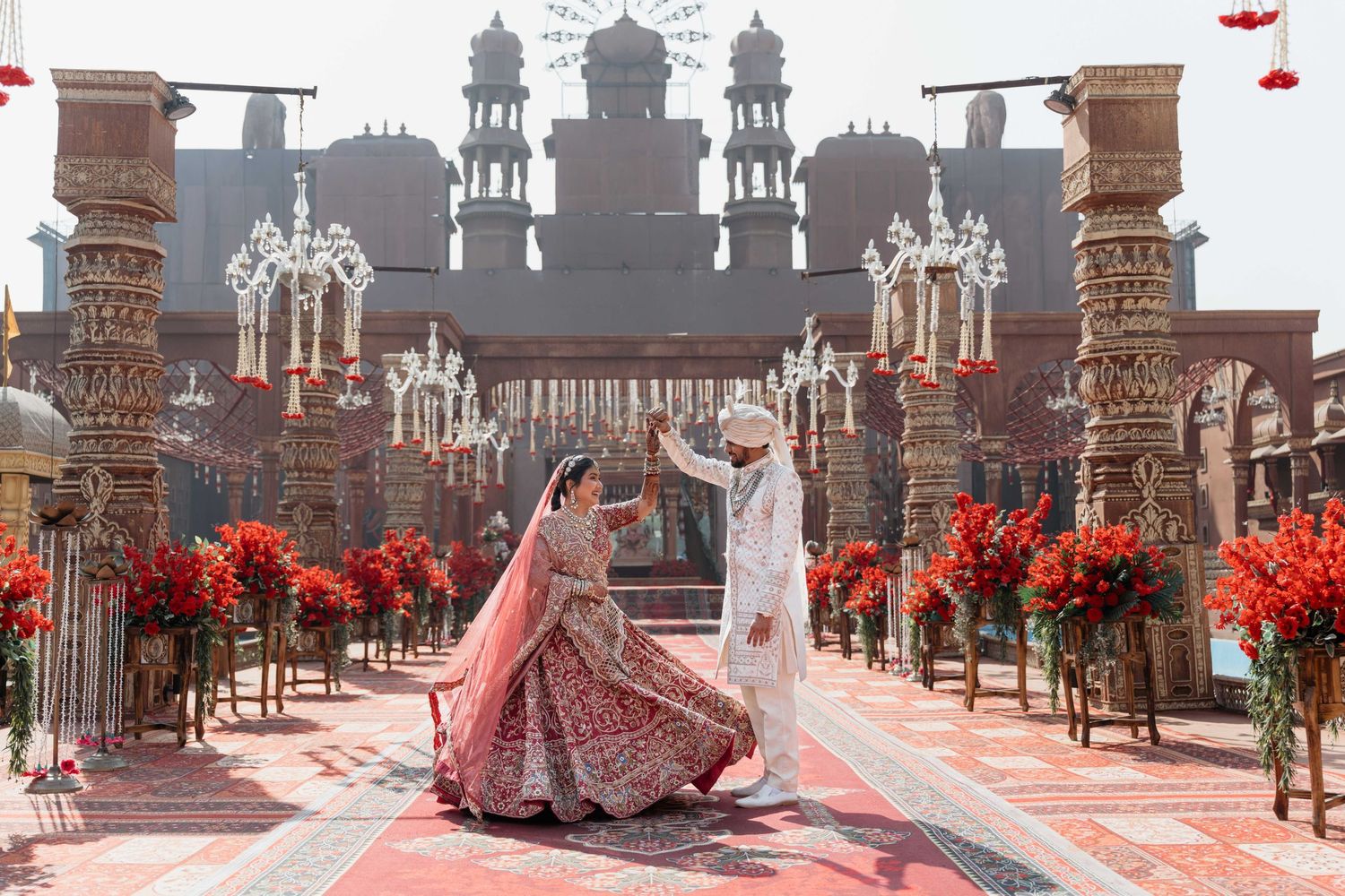 Bride and groom dancing in traditional Indian wedding attire at ornate temple venue with red floral decor.