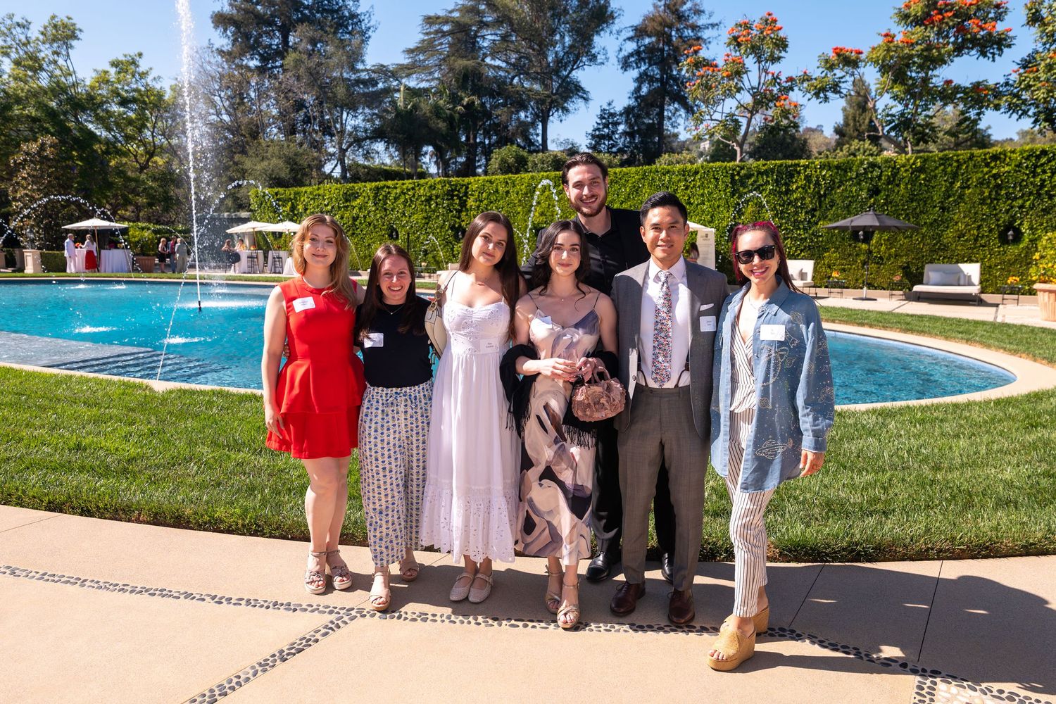 Group of seven friends posing together outdoors near a fountain at a formal garden event on a sunny day.