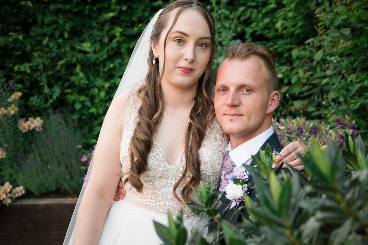 A bride and groom pose together in a lush garden setting, with the bride wearing a white lace gown and veil.